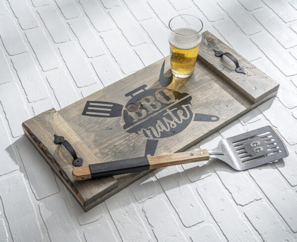 The Tray - BBQ, a wooden tray labeled "BBQ master," holds a glass of beer while a metal spatula with a wooden handle lies beside it on a white brick surface. - Spruced Studio Upscale Workshop