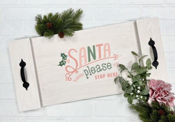 The "11x17 Tray - Santa Please Stop Here" is a white wooden tray with black handles, festive greenery, and a pink flower, featuring "Santa please stop here" in red and green lettering. - Spruced Studio Upscale Workshop