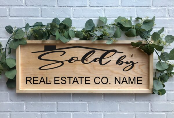 A 12x32 Sold By wooden sign with greenery reads "Sold by REAL ESTATE CO. NAME" against a white brick wall. - Spruced Studio Upscale Workshop