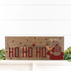 The 12x18 Santa Ho Ho Ho is a wooden sign featuring “HO HO HO” in red letters and a Santa Claus graphic, displayed against a white wall with green foliage at the base. - Spruced Studio Upscale Workshop