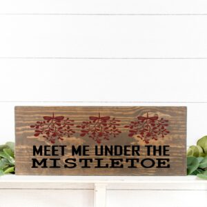 The 12x18 Meet Me Under the Mistletoe sign features three red mistletoe illustrations and the phrase "MEET ME UNDER THE MISTLETOE," displayed on greenery against a white wall. - Spruced Studio Upscale Workshop
