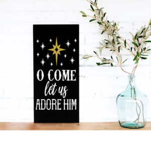 The 12x18 Oh Come Adore Him sign features white text and a gold star on black, displayed near a glass vase with olive branches on a wooden surface against a white brick wall. - Spruced Studio Upscale Workshop