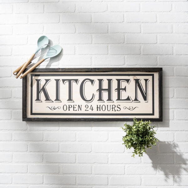 A 12x32 "Kitchen 24 hr - props" framed sign hangs on a white brick wall, surrounded by cooking utensils and a small potted plant. - Spruced Studio Upscale Workshop