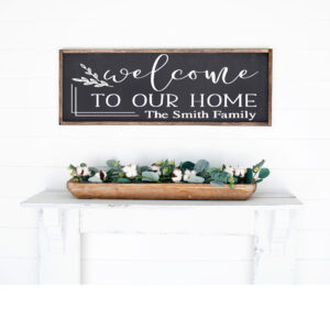 A 12x32 customizable "Welcome to our home" wooden sign hangs above a white shelf with a rectangular planter of greenery and cotton flowers. - Spruced Studio Upscale Workshop