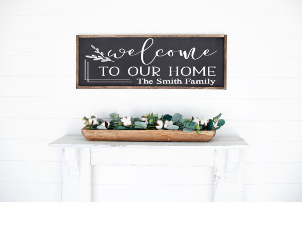 A 12x32 customizable "Welcome to our home" wooden sign hangs above a white shelf with a rectangular planter of greenery and cotton flowers. - Spruced Studio Upscale Workshop