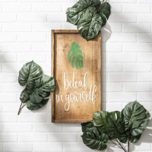 The 13x24 Beleaf - props 2 wooden sign, featuring a green leaf and the phrase "belak in yourself," is surrounded by green leaves and hangs on a white brick wall. - Spruced Studio Upscale Workshop