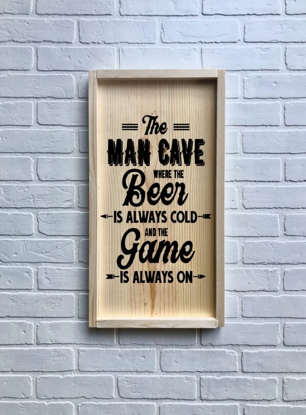 A 13x24 Cold Beer / Game On wooden framed sign hangs on a white brick wall, displaying: "The Man Cave where the beer is always cold and the game is always on. - Spruced Studio Upscale Workshop