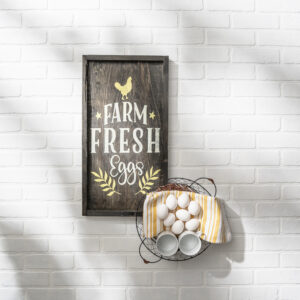 A 13x24 "Fresh Eggs" wooden sign hangs on a white brick wall above a wire basket filled with brown eggs, two white cups, and a yellow striped towel. - Spruced Studio Upscale Workshop