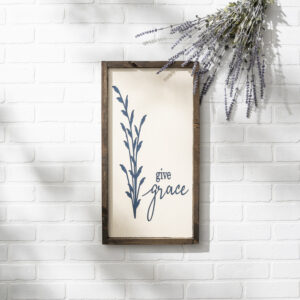 A 13x24 "Give Grace" framed sign featuring a blue botanical illustration hangs on a white brick wall beneath a bunch of lavender. - Spruced Studio Upscale Workshop