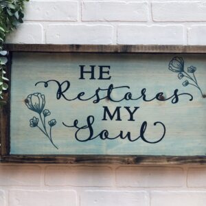 The 13x24 He Restores My Soul wooden sign features elegant cursive text and simple floral accents. Displayed on a white brick wall with greenery in the upper left corner, it brings a calming touch to any space. - Spruced Studio Upscale Workshop
