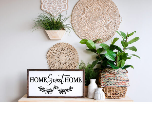 A 13x24 Home Sweet Home sign sits on a shelf with potted plants, wicker baskets, and white vases, set against a neutral wall decorated with woven hangings. - Spruced Studio Upscale Workshop