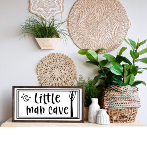 The 13x24 Little Man Cave (outdoors) sign is displayed on a shelf with woven baskets, greenery, and wall hangings in a neutral-toned room. - Spruced Studio Upscale Workshop