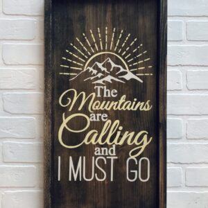 The 13x24 Mountains are Calling wooden sign features a mountain image and the quote "The Mountains are Calling and I Must Go," displayed on a white brick wall. - Spruced Studio Upscale Workshop