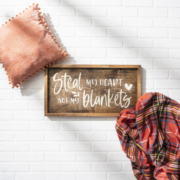The 13x24 Steal Heart Not Blanket sign is displayed on a white brick wall, styled with an orange pillow and a red plaid blanket as props. - Spruced Studio Upscale Workshop