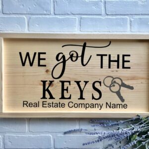 The 13x24 - We Got the Keys wooden sign features "We got the keys" and an image of keys, with "Real Estate Company Name" below, displayed against a white brick wall accented with lavender. - Spruced Studio Upscale Workshop