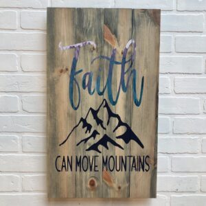 A 14x24 "Faith Can Move Mountains" wooden sign featuring a mountain design, displayed on a white brick wall. - Spruced Studio Upscale Workshop