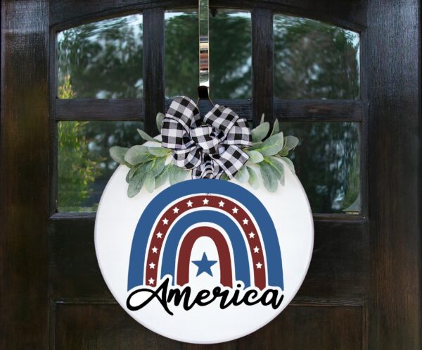 The America (rainbow) - Welcome Sign is a round door sign with a plaid bow, greenery, and a red, white, and blue rainbow with stars and stripes. "America" is displayed at the bottom. - Spruced Studio Upscale Workshop