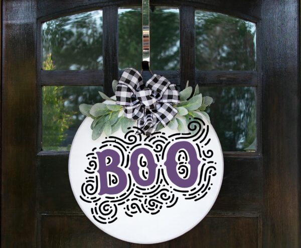 The "Boo" round white door sign features bold black letters, decorative swirls, greenery, and a black-and-white plaid bow, and is displayed on a dark wooden door. - Spruced Studio Upscale Workshop
