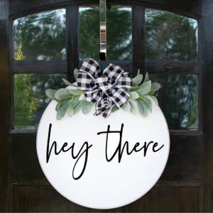 The "Hey There - Welcome Sign" features black script on a round white base, accented with green leaves and a black-and-white plaid bow, and is displayed on a dark wooden door. - Spruced Studio Upscale Workshop