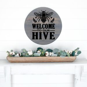 The "Welcome to Our Hive" round sign with a bee is displayed above a white shelf, where a wooden tray holds greenery and cotton stems. - Spruced Studio Upscale Workshop