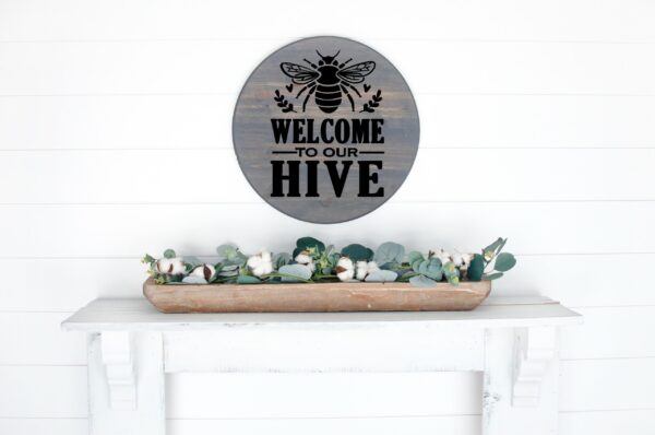 The "Welcome to Our Hive" round sign with a bee is displayed above a white shelf, where a wooden tray holds greenery and cotton stems. - Spruced Studio Upscale Workshop