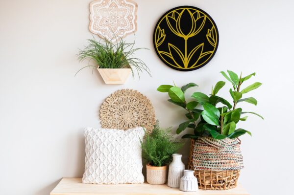 An assortment of houseplants in woven baskets and white vases sits on a light wooden surface; wall decor features two round woven pieces and an 18" Round - Flower art above. - Spruced Studio Upscale Workshop