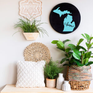 A wooden bench with potted plants, decorative pillows, woven decor, and the "18 Round - Michigan Home" wall art arranged against a white wall. - Spruced Studio Upscale Workshop