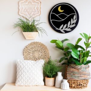 A decorative wall featuring the 18" Round - Moon & Mountains art, with plants in woven baskets, a white textured pillow, and ceramic vases on a light wooden surface. - Spruced Studio Upscale Workshop