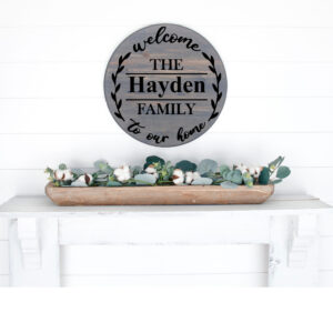 An 18" round wooden sign, "Welcome to Our Home" (customizable), hangs above a white mantel adorned with a wooden tray of greenery and cotton stems. - Spruced Studio Upscale Workshop