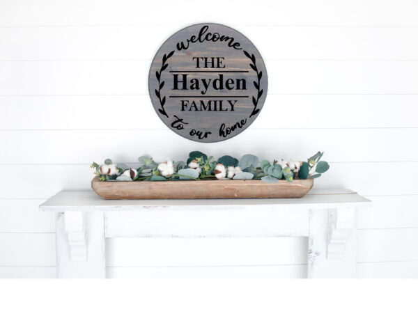 An 18" round wooden sign, "Welcome to Our Home" (customizable), hangs above a white mantel adorned with a wooden tray of greenery and cotton stems. - Spruced Studio Upscale Workshop