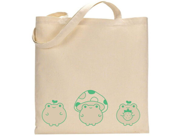 The "Three Frogs" beige tote bag features three green cartoon frogs—one holding a strawberry—and a large green mushroom at the center. - Spruced Studio Upscale Workshop