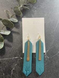 A pair of blue geometric earrings with gold accents and leaf engravings is displayed on a white card, set on a black marble surface with green leaves nearby. - Spruced Studio Upscale Workshop