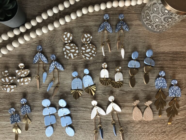 A collection of handmade polymer clay earrings in blue, white, and tan tones with various shapes and patterns displayed on a wooden surface. - Spruced Studio Upscale Workshop