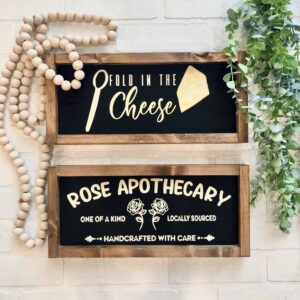 The 7x15 Schitt's Creek features two wooden-framed signs on a white brick wall: the top reads "Fold in the Cheese" with a cheese graphic, and the bottom reads "Rose Apothecary" accented by rose illustrations. - Spruced Studio Upscale Workshop