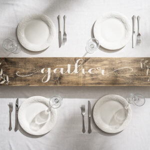 The 7x48 Tablerunner Gather is displayed on a white tablecloth, surrounded by four place settings with white plates, utensils, and glasses on a wooden board featuring "gather. - Spruced Studio Upscale Workshop