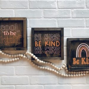 The 9x9 Be Kind framed sign features the phrase "Be Kind" with a rainbow design, displayed on a white brick wall and accented by a string of wooden beads. - Spruced Studio Upscale Workshop