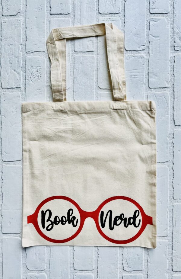 The Book Nerd (glasses) tote bag features red eyeglasses with "Book Nerd" inside the lenses, displayed on a white brick surface. - Spruced Studio Upscale Workshop