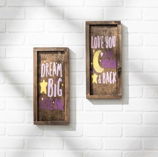 The 7x14 Dream Big wooden sign features "DREAM BIG little one" on a white brick wall beside a matching "LOVE YOU to the MOON & BACK" sign, both adorned with star and moon illustrations. - Spruced Studio Upscale Workshop