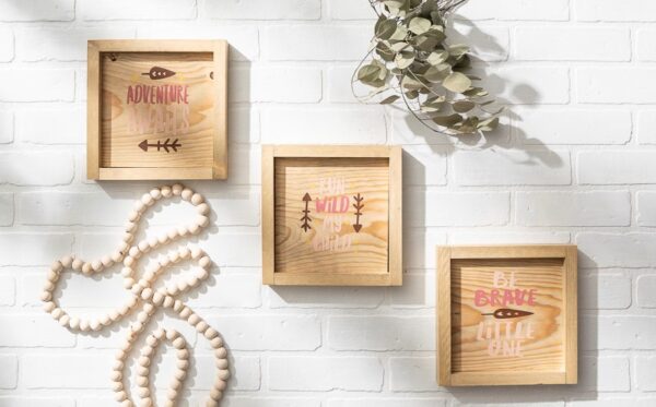 Three 9x9 wooden-framed signs—Run Wild, Adventure, and Be Brave—hang on a white brick wall beside a beaded garland and dried foliage. - Spruced Studio Upscale Workshop