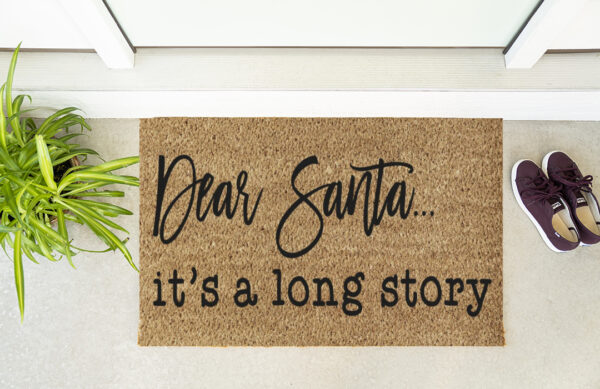The Santa It's a Long Story doormat sits by the door, accompanied by a nearby plant and a pair of maroon shoes. - Spruced Studio Upscale Workshop
