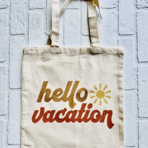 The "Hello Vacation" beige tote bag features the phrase and a yellow sun graphic on the front, shown against a white brick background. - Spruced Studio Upscale Workshop