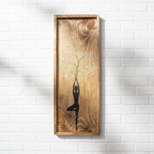 The 12x32 Yoga wooden wall art features a person’s silhouette in a yoga pose, arms raised to form a tree with green leaves, displayed against a white brick wall. - Spruced Studio Upscale Workshop
