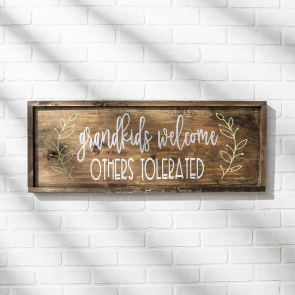 A 12x32 Grandkids Welcome wooden sign hangs on a white brick wall, with decorative leaves flanking the phrase: "grandkids welcome, OTHERS TOLERATED. - Spruced Studio Upscale Workshop