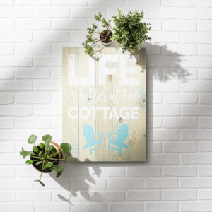 The 18x24 Cottage Life - props sign features "Life is better at the cottage" with two blue chairs, mounted on a white brick wall accented by two hanging potted plants. - Spruced Studio Upscale Workshop