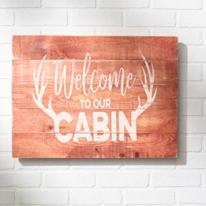 The 18x24 Welcome Cabin sign features white "Welcome to our cabin" text with antler graphics on wood, shown mounted on a white brick wall. - Spruced Studio Upscale Workshop