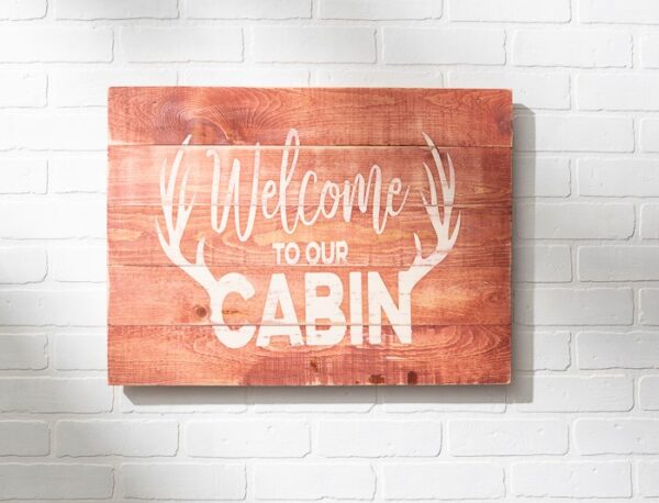 The 18x24 Welcome Cabin sign features white "Welcome to our cabin" text with antler graphics on wood, shown mounted on a white brick wall. - Spruced Studio Upscale Workshop