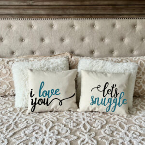 The "I Love You + Let's Snuggle" decorative pillows are displayed on a neatly made bed with a patterned beige comforter and a tufted headboard. - Spruced Studio Upscale Workshop