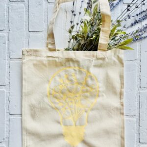 The Lightbulb & Wildflowers beige tote bag, featuring a yellow lightbulb and floral design, filled with green and purple foliage, rests on a white textured surface. - Spruced Studio Upscale Workshop