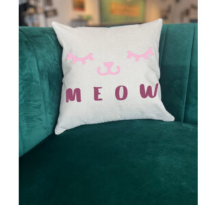 A Meow (cat face) pillow featuring a pink cat face and "MEOW" in bold letters rests on a green velvet sofa. - Spruced Studio Upscale Workshop