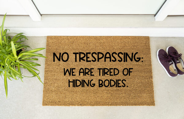 The "No Trespassing We Are Tired of Hiding Bodies" doormat is shown by a door, next to a plant and a pair of shoes. - Spruced Studio Upscale Workshop